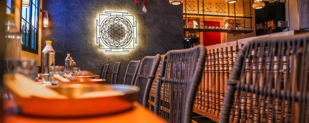 Elegant restaurant interior with a long wood table, wicker chairs, and a large illuminated geometric mural on a dark stone wall.
