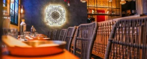 Elegant restaurant interior with a long wood table, wicker chairs, and a large illuminated geometric mural on a dark stone wall.