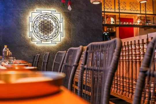 Elegant restaurant interior with a long wood table, wicker chairs, and a large illuminated geometric mural on a dark stone wall.