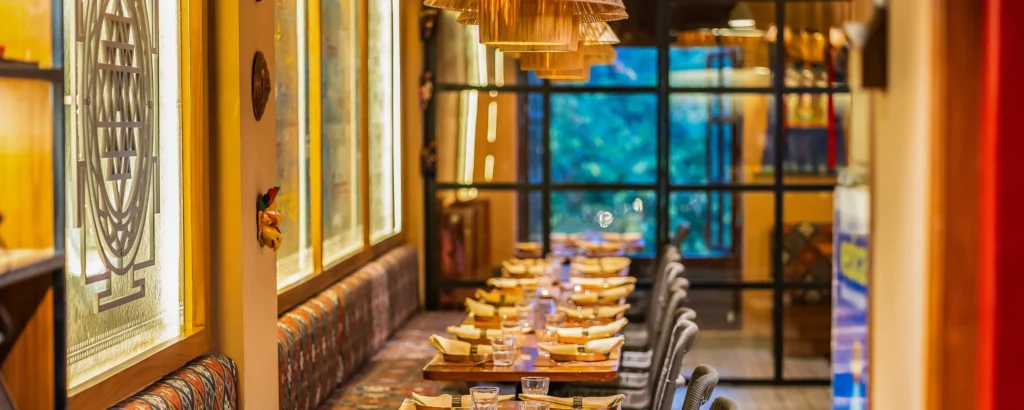 Cozy restaurant interior with a long row of wooden tables set for dinner, glassware and napkins, warm pendant lights overhead, and a patterned bench along the left wall.