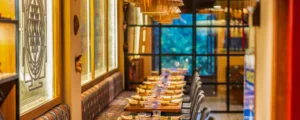 Cozy restaurant interior with a long row of wooden tables set for dinner, glassware and napkins, warm pendant lights overhead, and a patterned bench along the left wall.