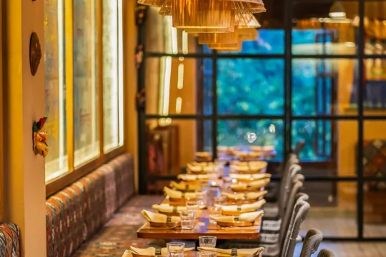 Cozy restaurant interior with a long row of wooden tables set for dinner, glassware and napkins, warm pendant lights overhead, and a patterned bench along the left wall.